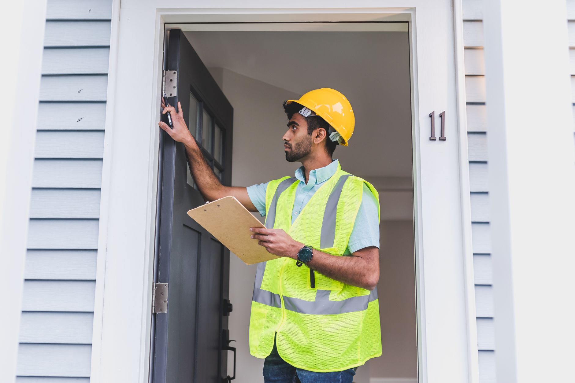 A contractor in a hard hat and safety vest inspecting a doorframe with a clipboard during a site visit.
