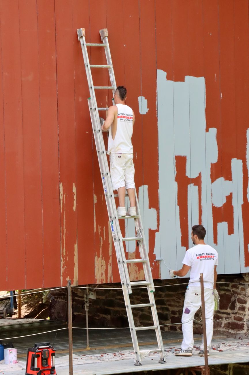 Two painters in white overalls work on ladders to repaint the wooden siding of a house.