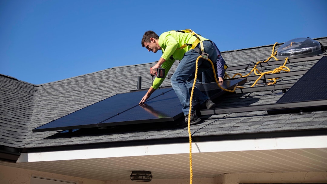 Installer wearing a safety harness kneeling on a shingle roof while positioning a solar panel next to a partially completed array